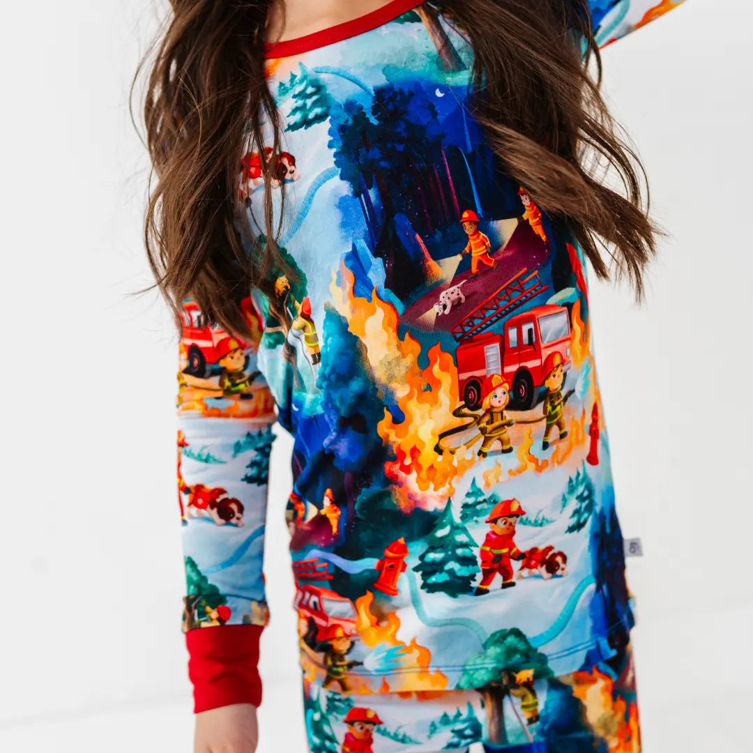 Close-up of a child wearing a pajama set with a print of firefighters, red fire trucks, and flames in a snowy forest scene.
